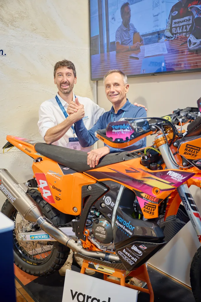 Dos personas se dan la mano junto a la moto de rally con el logotipo de Varador 2000 en el stand del Monaco Yacht Show